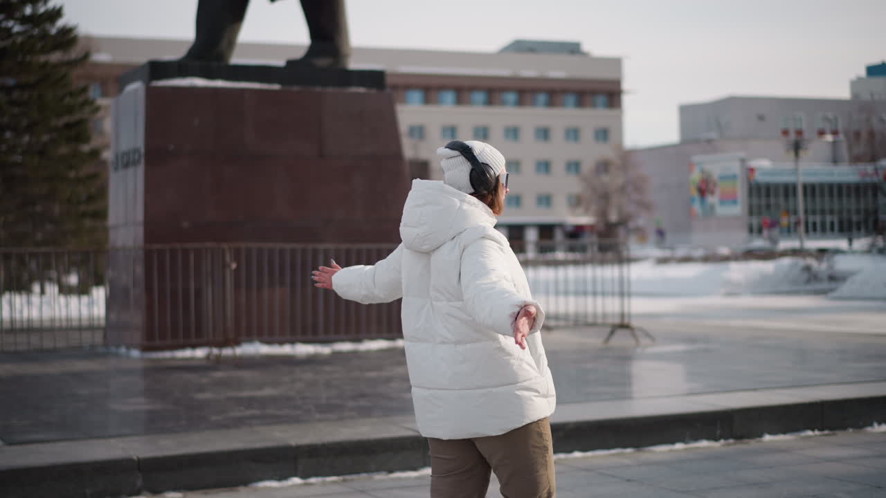 Round view of student wearing white coat and headset dancing on tiled plaza in front of school near statue with snow and trees under bright winter daylight and passing pedestrians