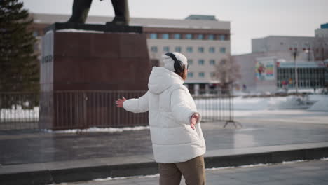 round view of student wearing white coat and headset dancing on tiled plaza in front of school near statue with snow and trees under bright winter daylight and passing pedestrians