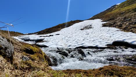 Schmelzender-Schnee-Im-Frühling-Fließt-In-Richtung-Sysen-Dam-Wasserreservoir-Für-Das-Wasserkraftwerk-Hardangervidda-Norwegen---Statische-Nahaufnahme-Des-Stroms-In-Der-Nähe-Des-Schneeflecks-Neben-Gelbem-Verwelktem-Gras