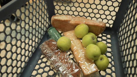 a lady in a black coat and black boots placing bananas into a shopping trolley containing a pack of biscuits, a small juice, a loaf of bread, and seven apples