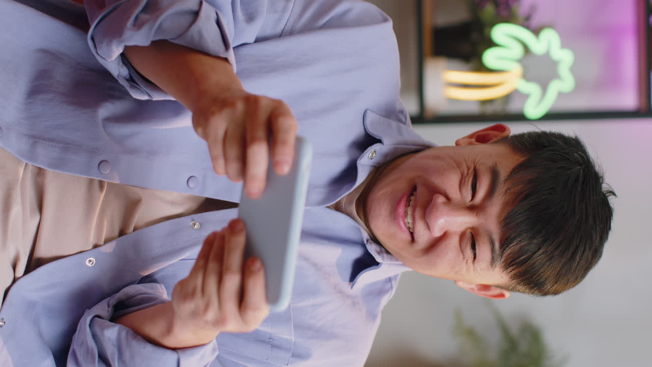 걱정스러운 아시아의 잘생긴 남자가 집에서 소파에서 스마트폰에서 레이싱 또는 슈팅 비디오 게임을 하고 있습니다.