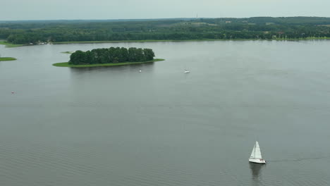 Aerial view of a small island in the middle of a calm lake with a sailboat cruising nearby - Jeziorak lake