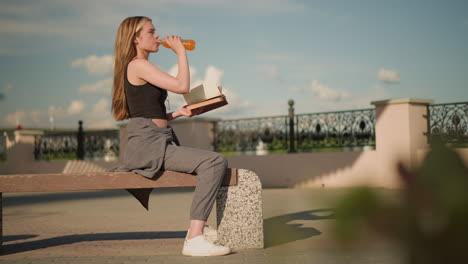 woman seated outdoors, holding book in one hand while sipping from juice bottle with the other, she continues reading while casting shadow on floor in warm daylight, with background fence