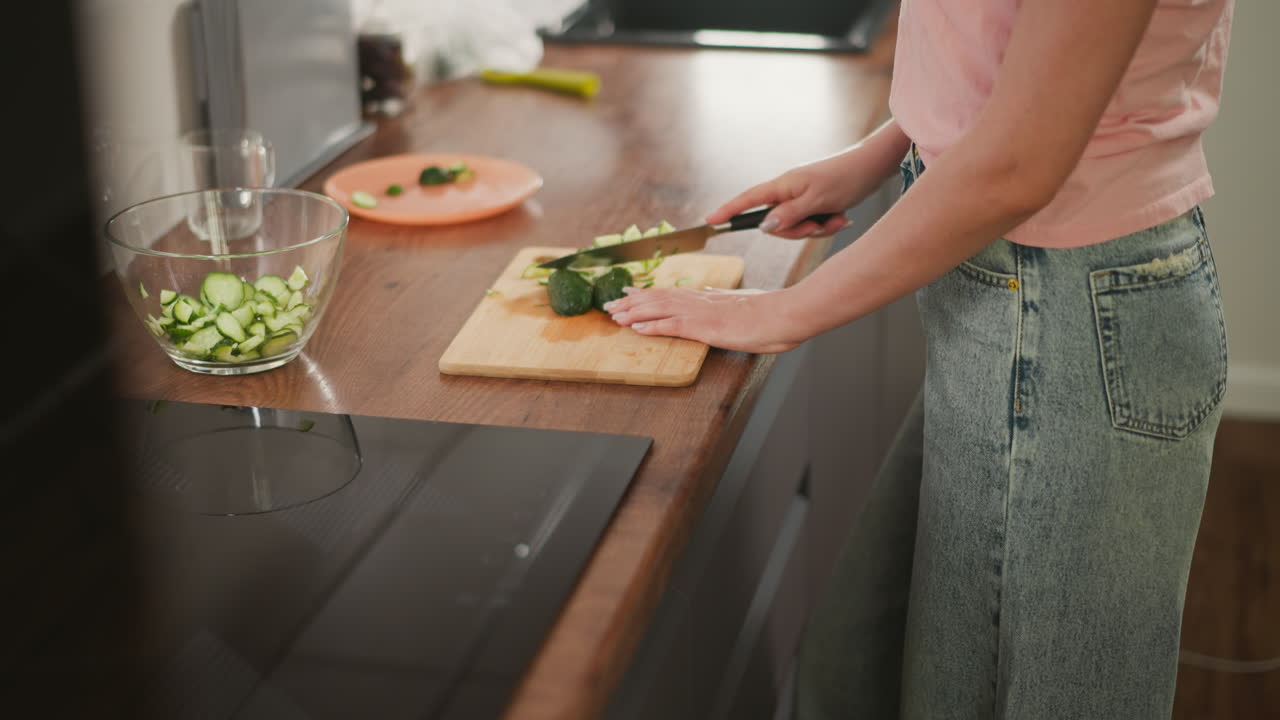 Side view of person wearing casual clothes slicing fresh cucumber on wooden cutting board beside glass bowl with sliced vegetables on kitchen countertop in clean and modern home cooking environment