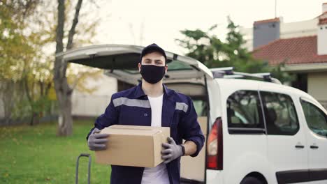 handsome courier takes cardboard box package out of delivery van walks through private sector. courier in mask, cap and gloves