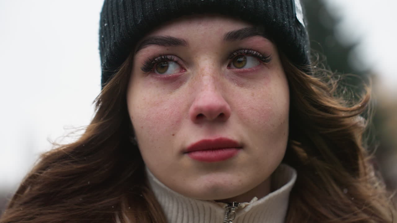 Close up of thoughtful girl in black cap and brown jacket walking with serious expression and wind-blown hair against blurred city background during cold quiet day with overcast sky