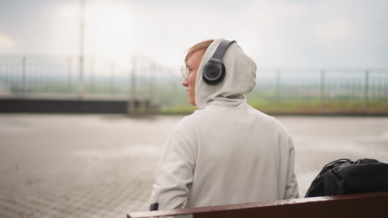 흐린 하늘 아래 안개가 자욱한 수평선과 텅 빈 산책로를 바라보며, 후드티를 입고 헤드폰을 낀 채 물가의 벤치에 앉아 책장을 넘기는 외로운 백인 여행자의 사색적인 휴식