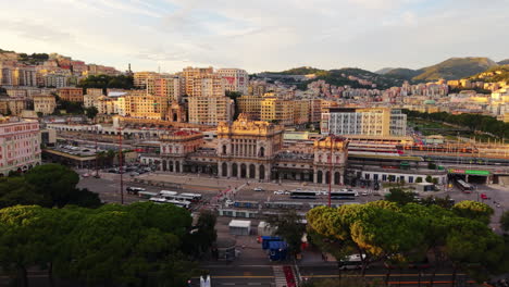 drone flying forward above the tree-lined street leading to genoa train station at sunset, with modern and historic buildings, bus stops, and mountains in the distance