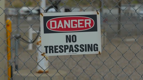 a close up shot of a danger and no trespassing sign attached to a chain-linked fence with cars driving in the background