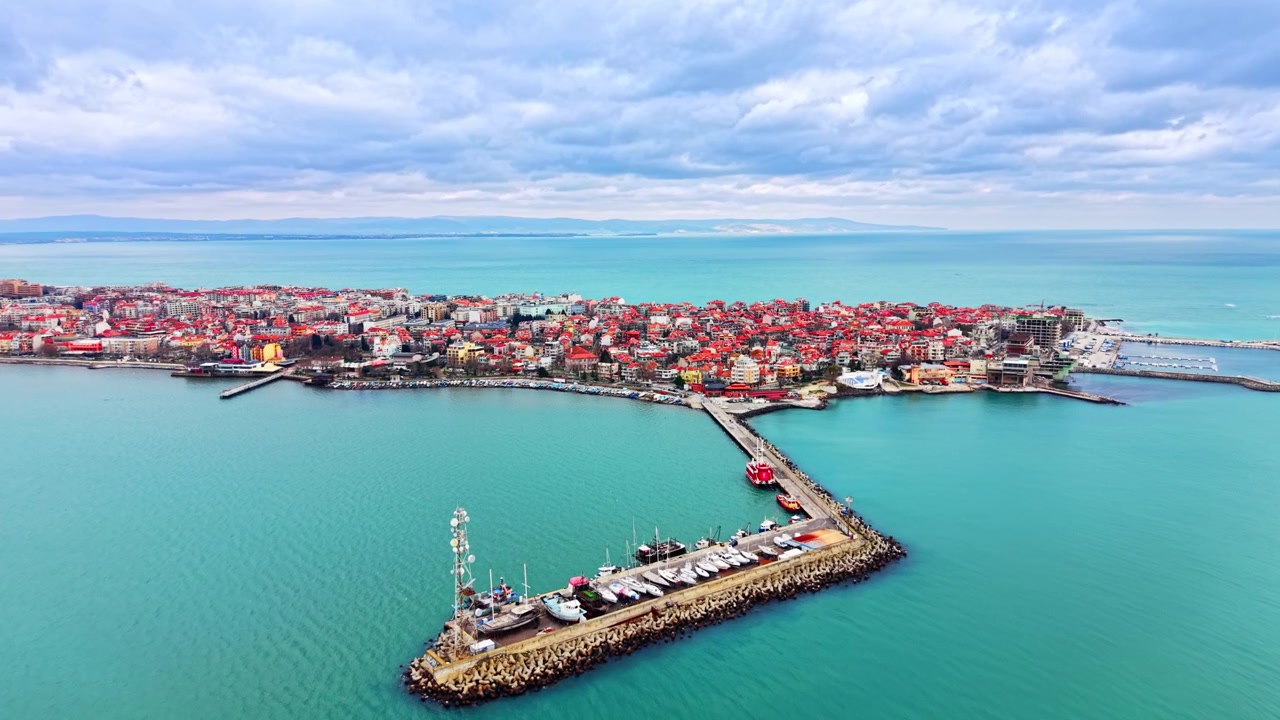 Flygfoto över halvöstad med stenpir, Bulgarien. En smal stenig pir håller en förtöjd yacht och ett fiskefartyg bredvid en tätbebyggd kustbosättning