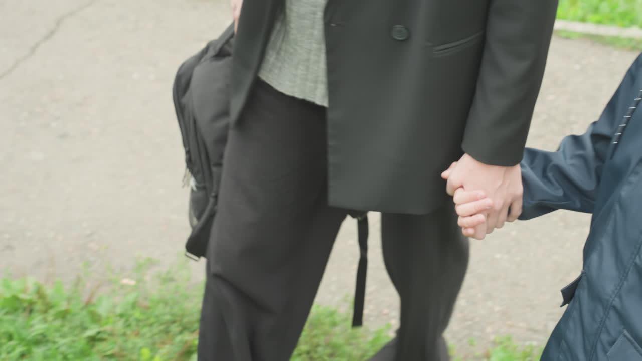 保護者が子供に付き添っている。大人と子供はバックパックを背負い、自信を持って一緒に舗道を歩いている。大人が幼い子供を、確かな足取りで安全に歩道へと導いている。