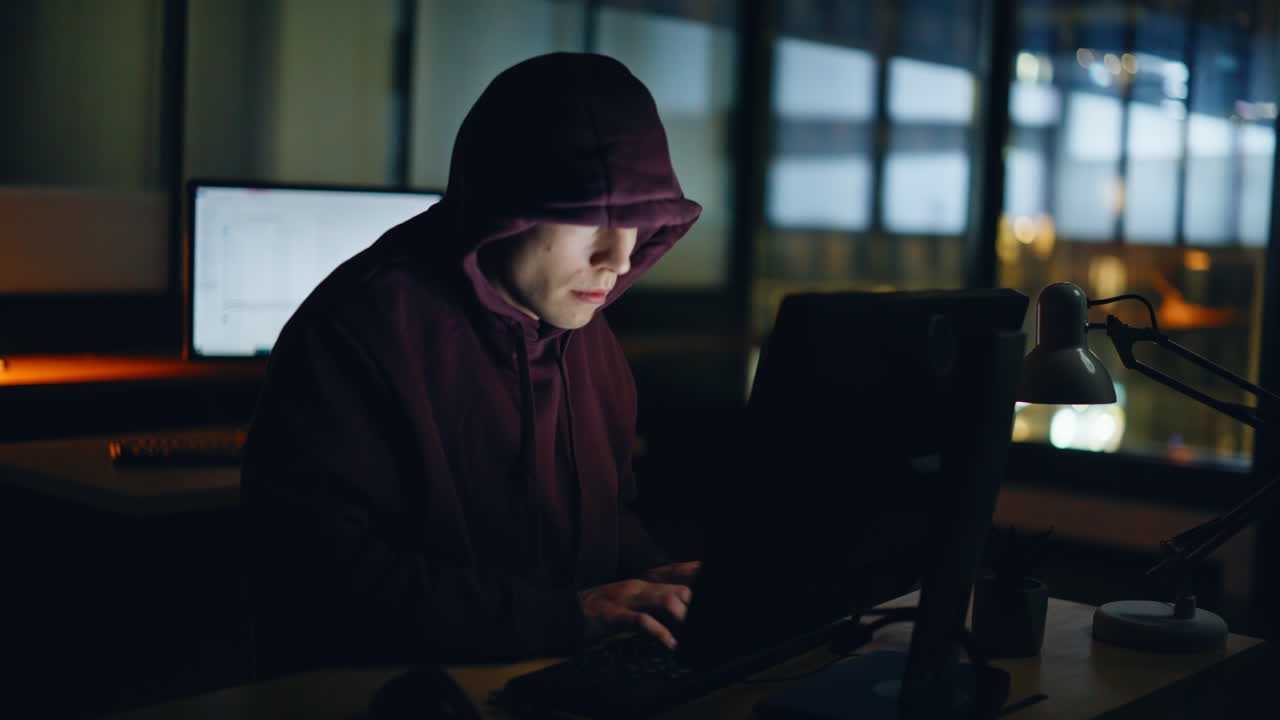 Hætteklædt hacker bryder ind i dataservere i et lukket rum om natten. Mand udfører cyberangreb.