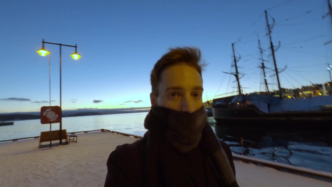 Oslo, Norveç'teki Aker Brygge'de karla kaplı liman boyunca yürüyen, atkıya sarılmış bir adamın soğuk bir kış şafağında çekilmiş yakın çekim selfie tarzı görüntüsü.