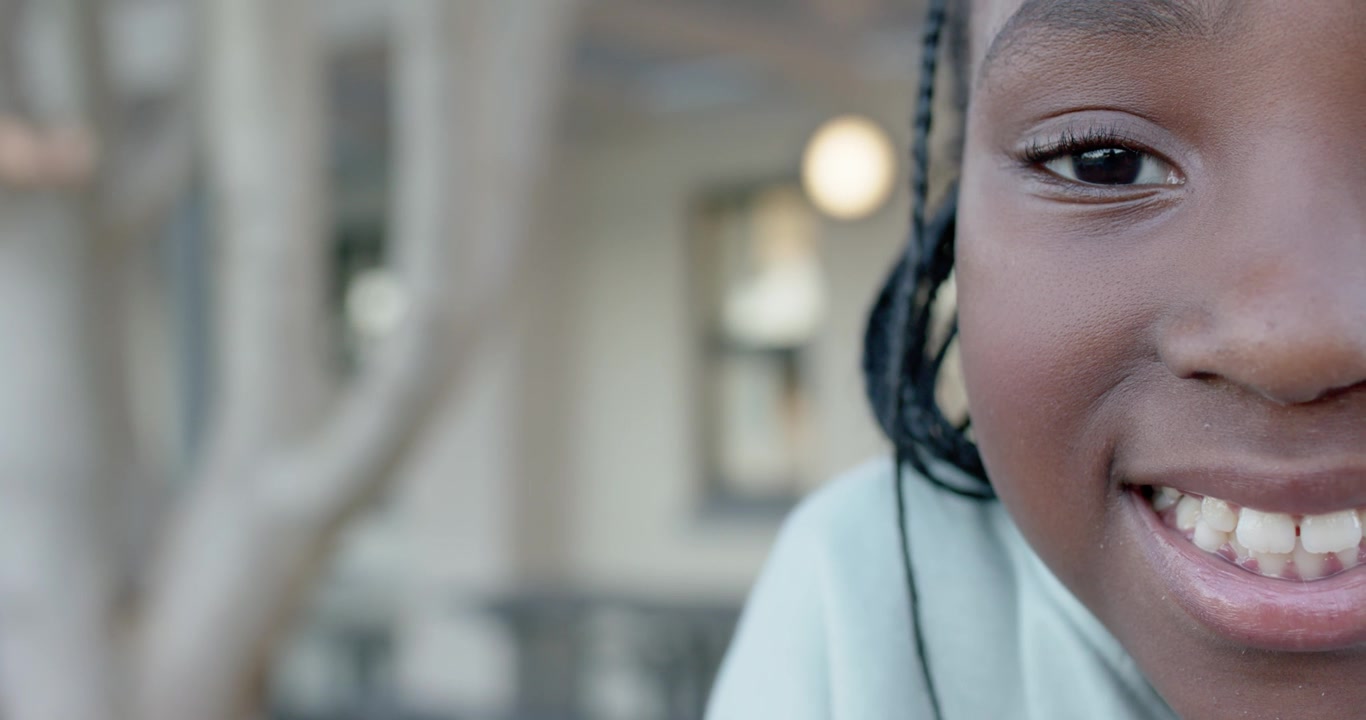 Bambino africano che si avvicina alla telecamera sorridendo, strizzando gli occhi su un portico con trecce, albero, spazio per il testo