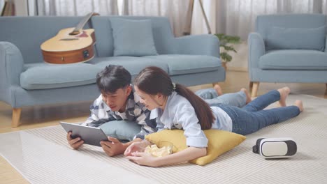 full body of asian teen couple using tablet while lying on carpet on the floor at home. watching movie on tablet with snacks, speaking, and enjoying time together