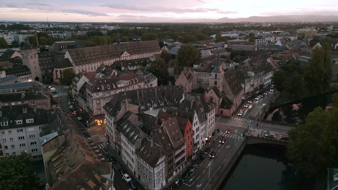 Atmospheric twilight aerial view over Strasbourg’s winding streets and rooftops, with soft evening lights illuminating the historic architecture and calm riverbanks as the city transitions into night