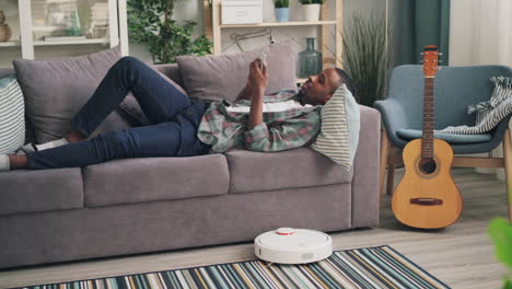 man relaxing on a couch with a smartphone and a robotic vacuum cleaner in the living room.