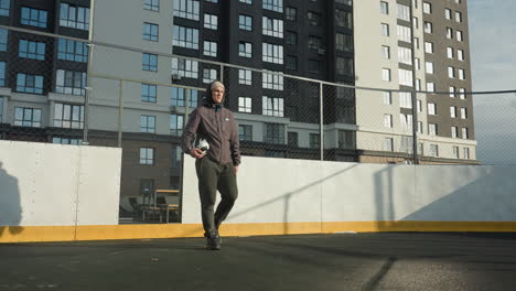 footballer in hoodie and beanie holding ball in right hand walks along football pitch with goal post, tall residential building in background, and sunlight casting shadow on ground