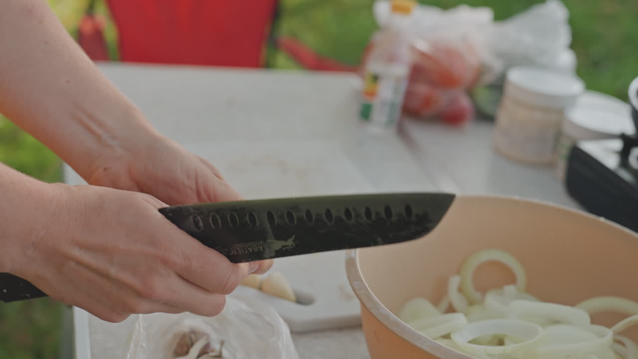 Donna che affetta finemente la cipolla in una ciotola al tavolo da campeggio all'aperto, primo piano su pelapatate e coltello, mani ferme che formano anelli precisi, luce solare soffusa e sfondo verde, ritmo concentrato per la preparazione dello stufato da campeggio.
