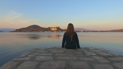 Back-view-of-child-girl-sitting-on-pier-edge-looks-at-Angera-castle-on-Maggiore-lake-in-Italy-at-sunset