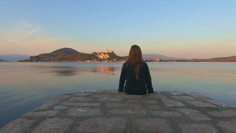 back view of child girl sitting on pier edge looks at angera castle on maggiore lake in italy at sunset