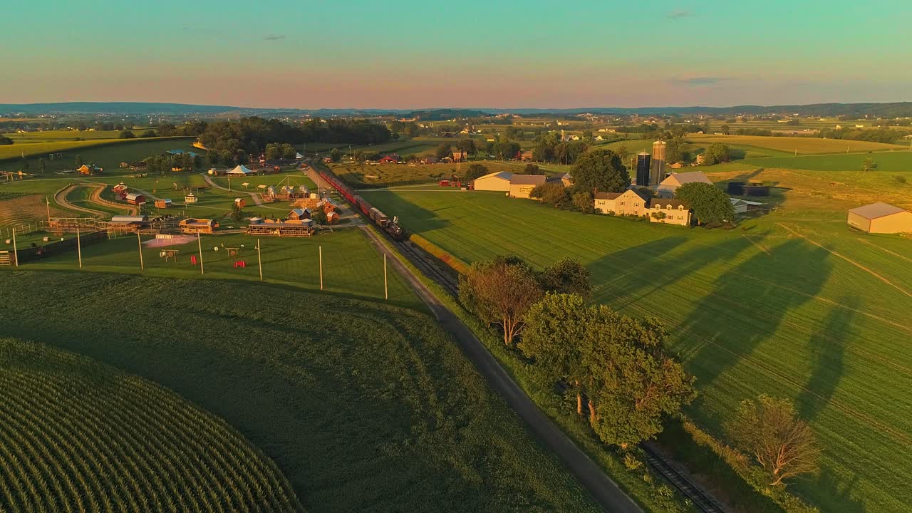 una vista aérea de un tren de pasajeros de vapor que se acerca pasando por un laberinto de maíz y campos de maíz durante la hora dorada en un día de verano