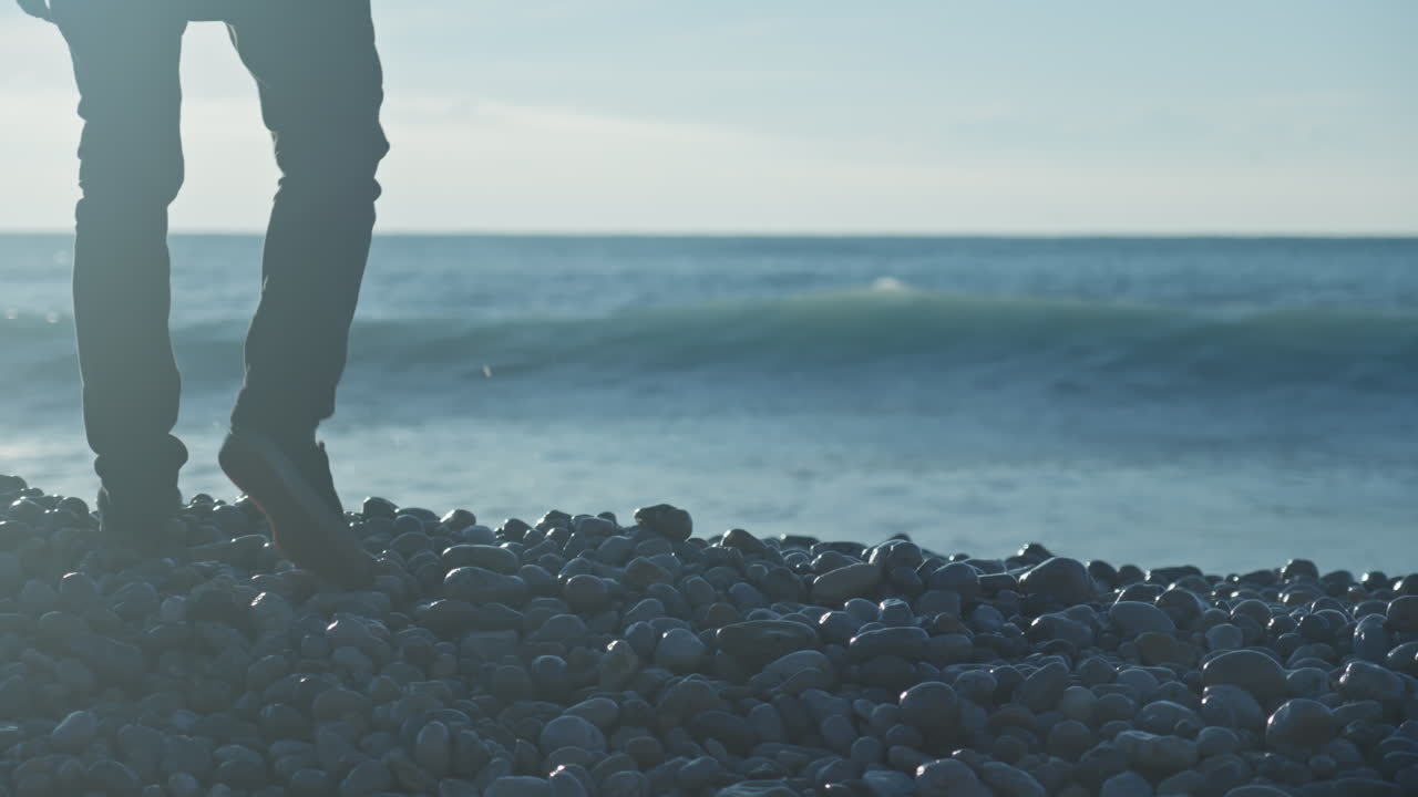 남자는 화창한 아침 일찍 해변의 바위 능선 위로 걸어가고, 파도가 부서지고 배경에서 튀는 동안 그는 마침내 바다에 돌을 던지고 있습니다.