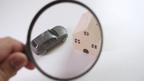 using a magnifying glass, an individual is showing investment possibilities in assets and real estate as represented by a toy car and a wooden toy house