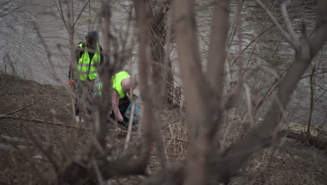 volunteers in reflective safety vests cleaning polluted riverbank, collecting plastic bottles and trash into bags, walking along muddy shoreline surrounded by bare trees during cold season