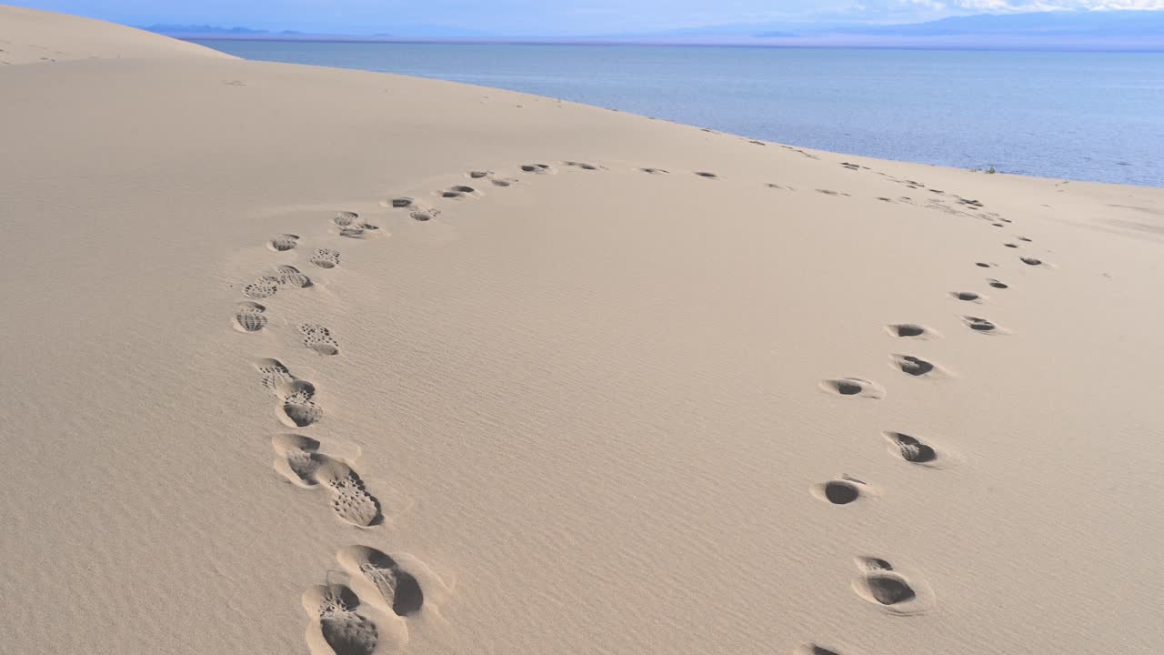 A scene of stunning contrast where the Durgun Nuur desert meets a vast lake. Footprints mark a solitary path across the sand dunes towards the water