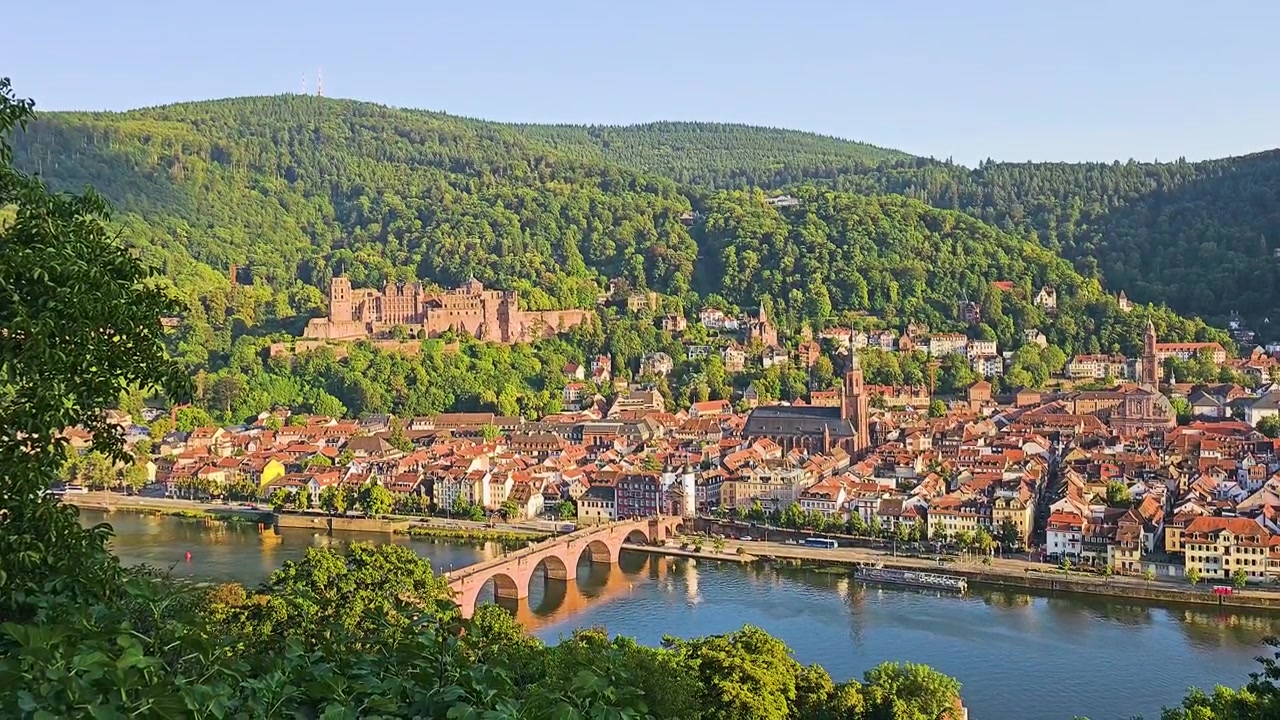 Panoramica della città di Heidelberg, Germania, con rovine del castello, il Ponte Vecchio, il fiume Neckar, tetti rossi, campanili e colline boscose che risplendono nella calda luce estiva da un punto panoramico.