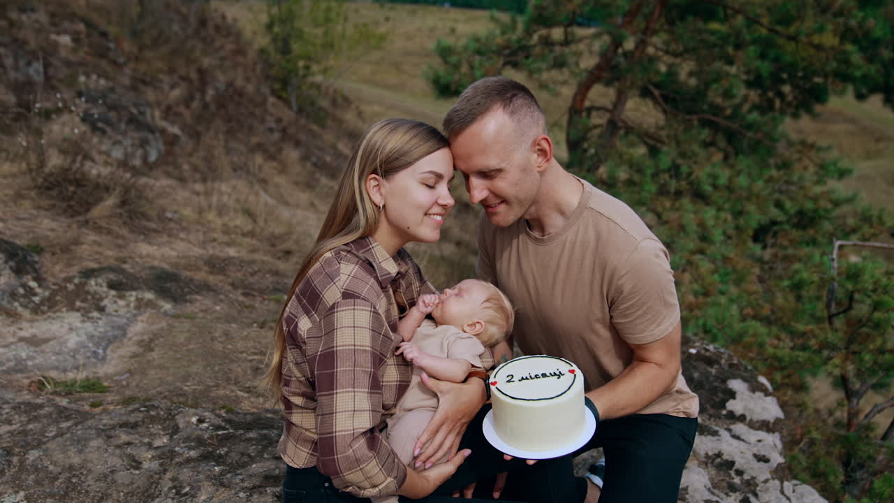 Celebrating two month of baby life. Happy parents hold an infant and cake sitting in the nature.