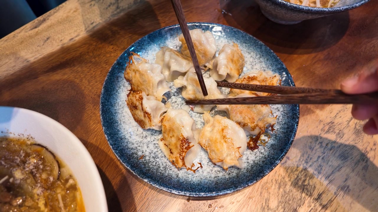 Close-up of wooden chopsticks picking up golden pan-fried dumplings at a restaurant in Bangkok.
