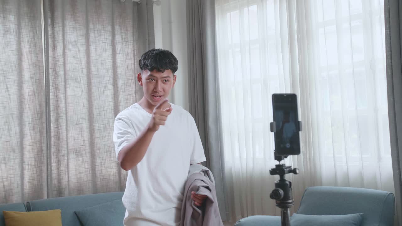 Ragazzo asiatico che mostra un trucco di sparizione con la fotocamera del cellulare mentre registra contenuti video sullo smartphone per i social network in una stanza accogliente