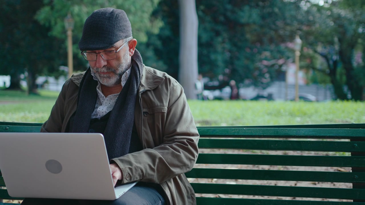 Elderly Man Sitting on Park Bench and Typing on Laptop