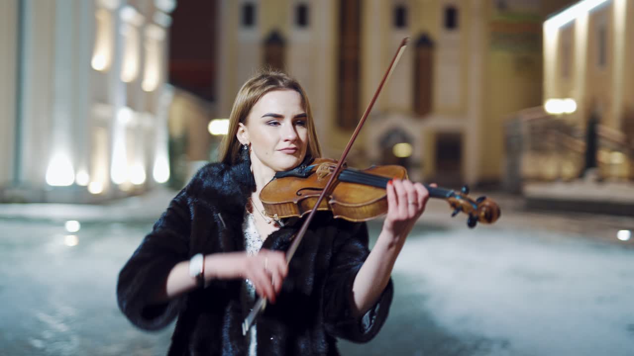 A student of musical faculty in an evening dress and coat is playing a famous music on violin on the background of the monument and city buildings behind her. Close-up