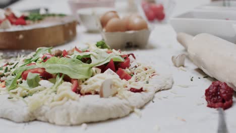 Close-up-of-cheese-and-vegetables-on-pizza-dough-with-kitchen-equipment-on-counter,-in-slow-motion