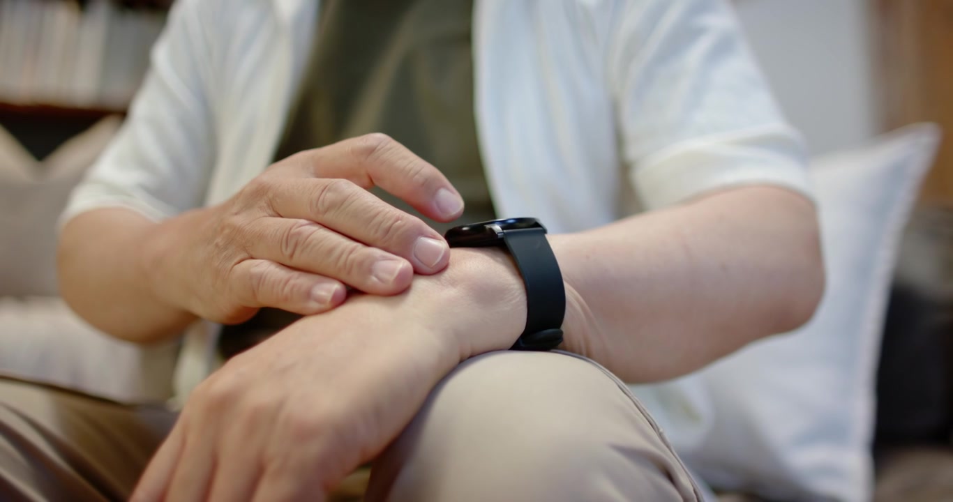 Uomo anziano asiatico appoggia l'avambraccio sinistro con le dita vicino allo smartwatch, toccando e scorrendo sul divano.
