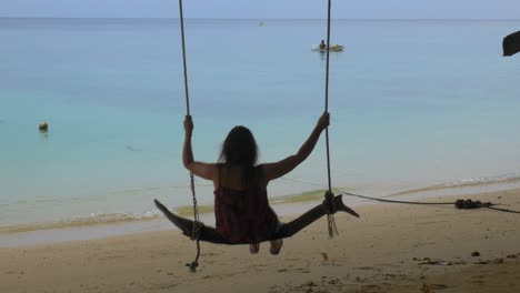 A-brunette-woman-swings-on-a-wooden-swing-on-a-tropical-beach,-shot-from-behind