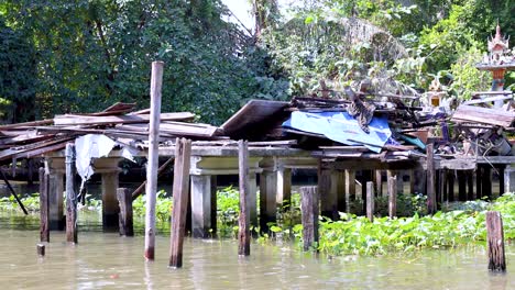 a dilapidated wooden house stands by a canal in bangkok, surrounded by lush greenery and reflecting a serene yet neglected atmosphere