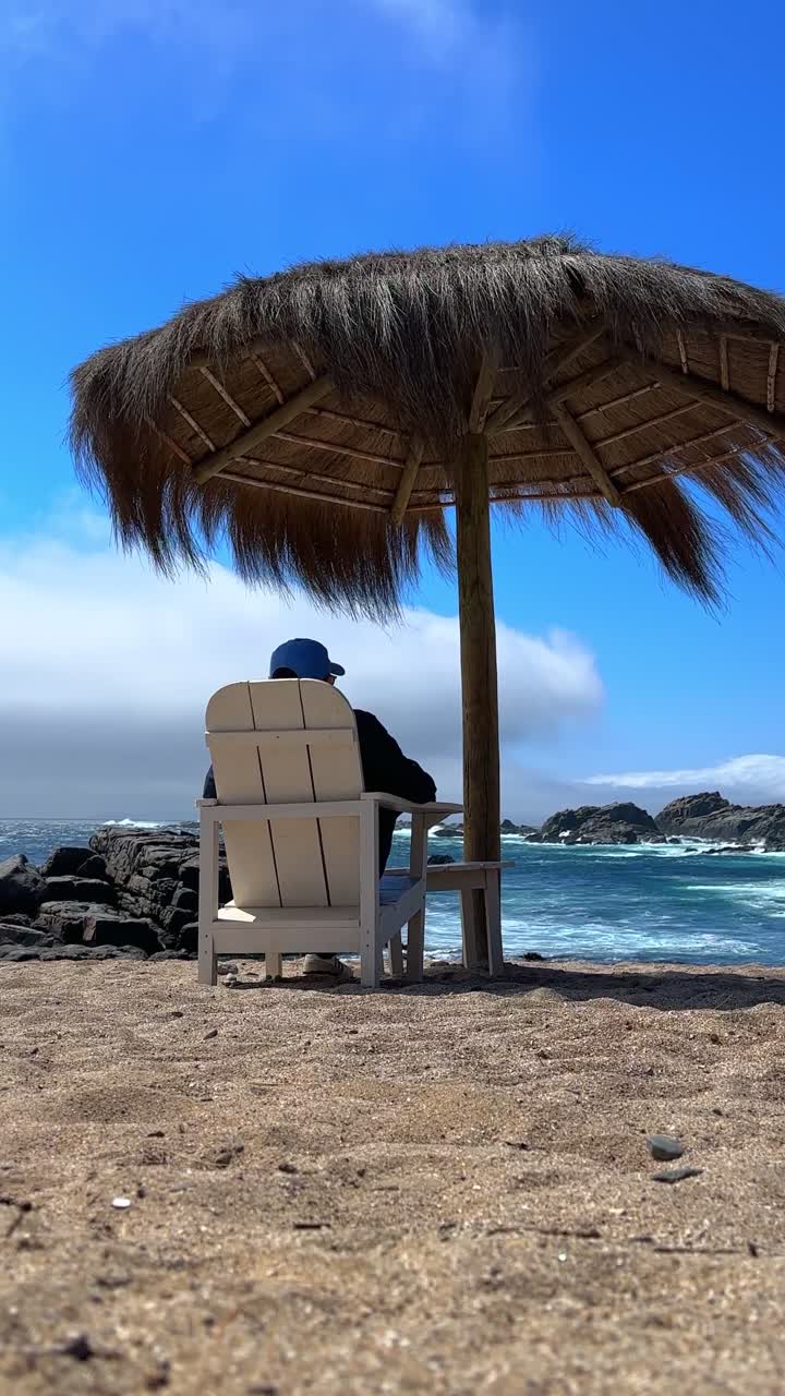 Video verticale di un giovane uomo seduto su una sedia di legno sotto una palma con tetto di paglia sulla spiaggia di Punta Pite, sulla costa centrale del Cile