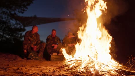 a large bonfire flame, behind it military men in camouflage uniforms are sitting, talking and warming themselves at night by the hearth