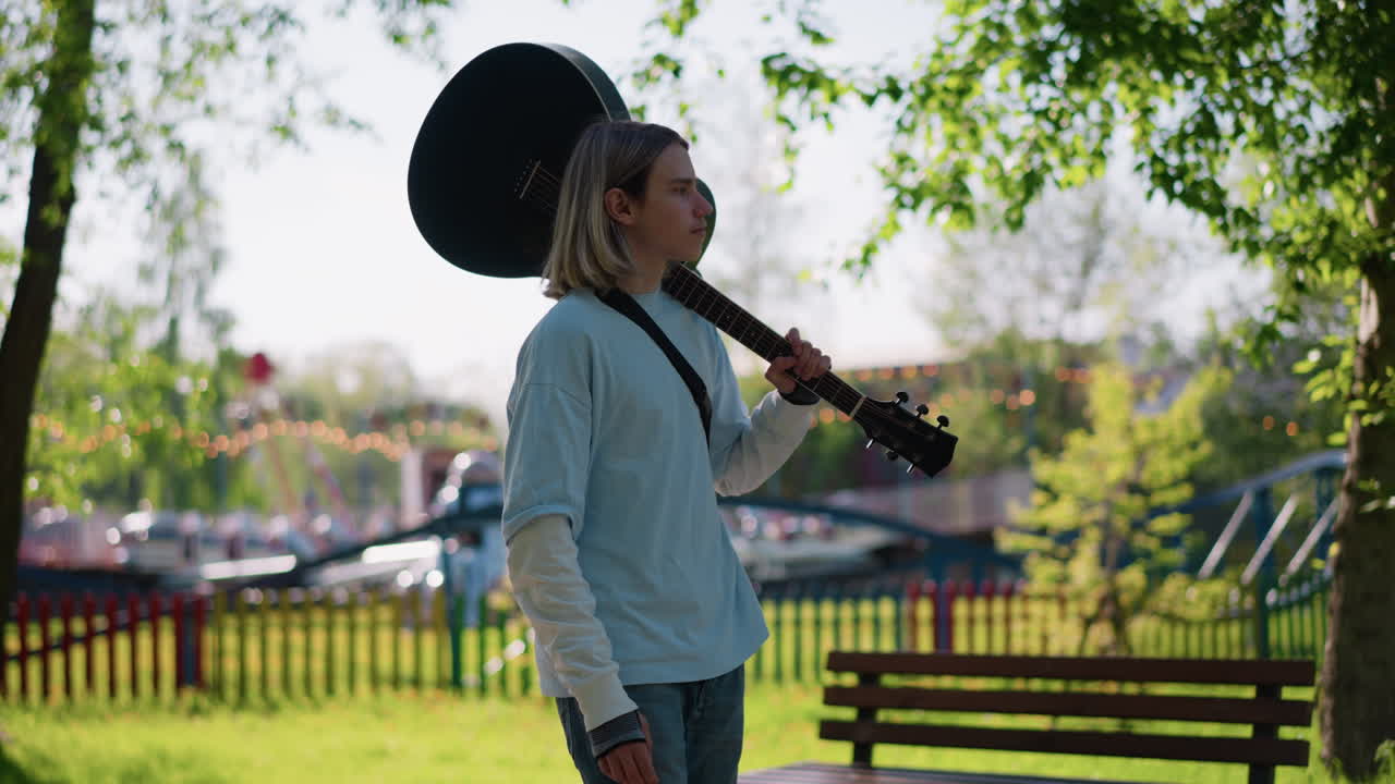 Un chitarrista che cammina in un parco assolato con una chitarra acustica sulla spalla, jeans e felpa casual, alberi e staccionata di legno sullo sfondo, luce estiva soffusa, atmosfera rilassata e contemplativa, ambientazione in un giardino urbano