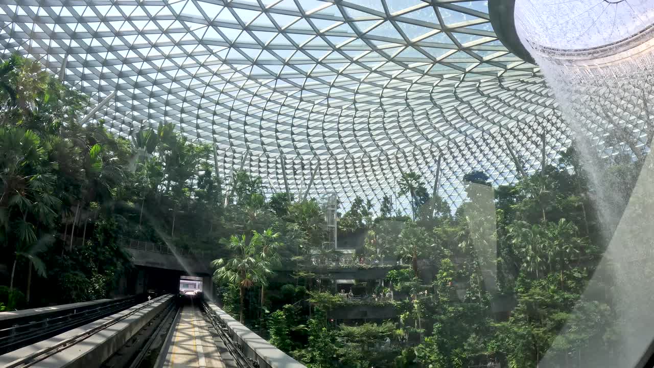 Automated train moves toward large indoor waterfall under glass dome with lush greenery, daylight illumination