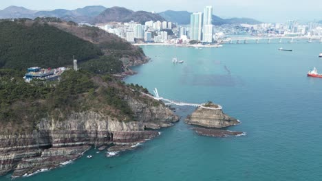 drone aerial view in south korea countryside flying over the clear blue sea of busan next to a green mountain, ships and city in the background sunny day