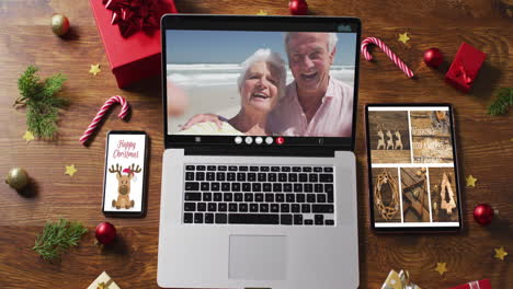 caucasian senior couple waving on video call on laptop, with smartphone, tablet and decorations