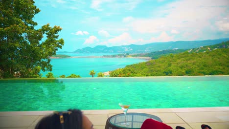 a woman with a sexy evening dress on a trip sitting on a sofa by the pool drinking and looking at the sea