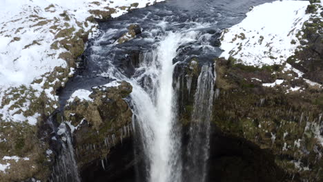 AERIAL:-Over-Waterfall-in-Snow,-Ice-Canyon-in-Iceland-Green-Grass,-Cloudy