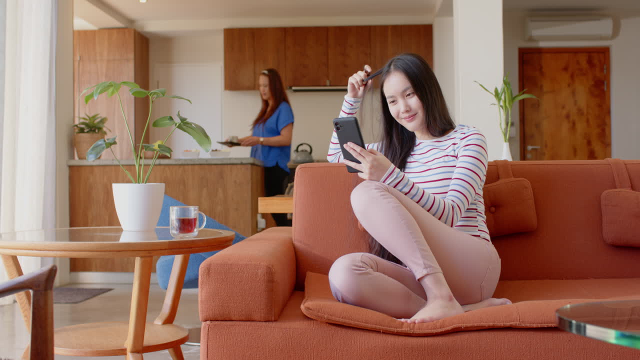 Using smartphone, young asian teenage girl sitting on couch while asian mother cooking in kitchen