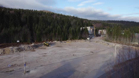 Volando-Sobre-Un-Terreno-Preparado-Para-Un-Sitio-De-Construcción-Con-Una-Excavadora-Y-Un-Bosque-Al-Final-Del-Sitio